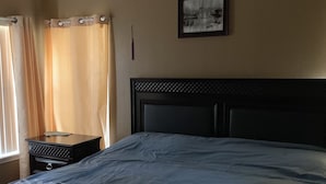 Un dormitorio con cama, mesita de noche, un cuadro en la pared y ventana con cortinas.