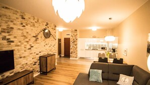 A modern living room with a brick wall, a flat-screen TV, a dark sofa, and a kitchen in the background. - Baltic Apartments - Apartament Aurora (Swinoujscie)