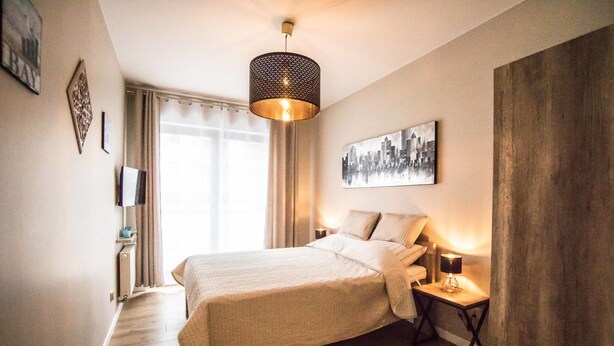 A hotel room with a large bed, a bedside table with a lamp, a cityscape painting on the wall, and a window with curtains. - Baltic Apartments - Apartament Aurora (Swinoujscie)