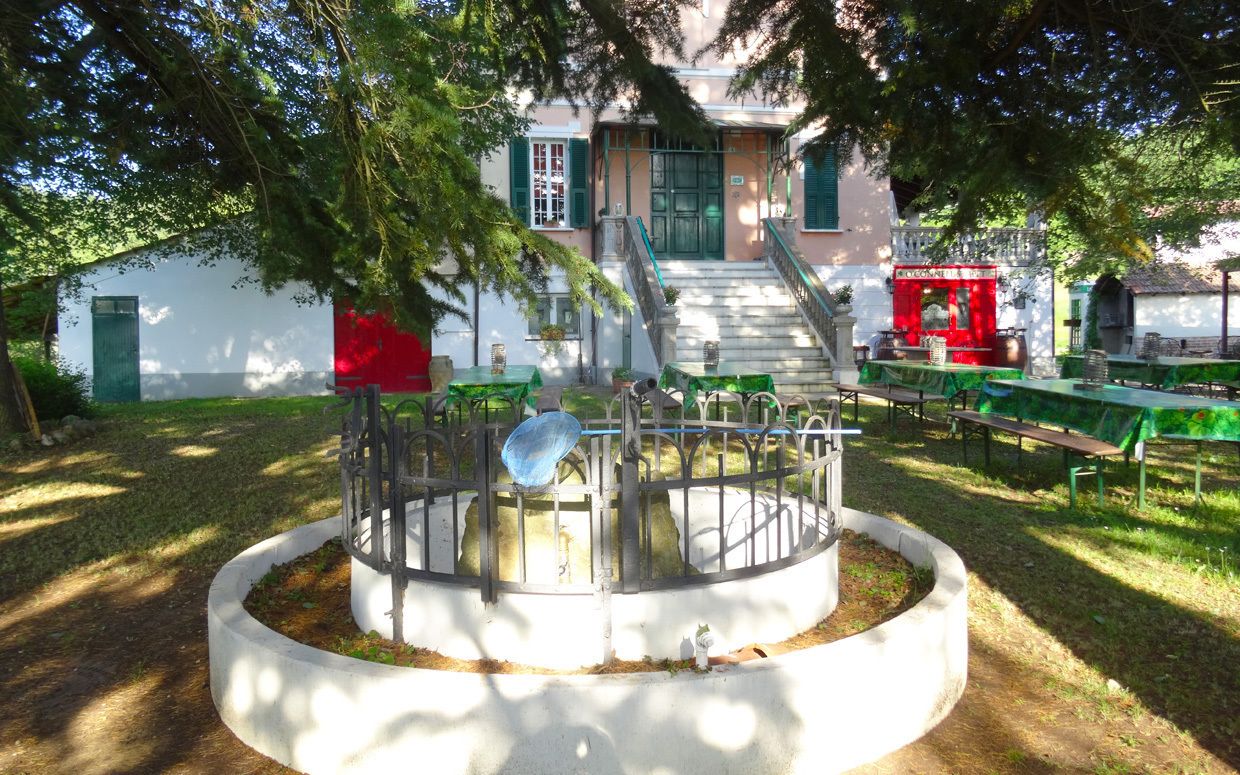 Un patio con una estructura circular central, una escalera que lleva a un edificio con contraventanas verdes y mobiliario de exterior.