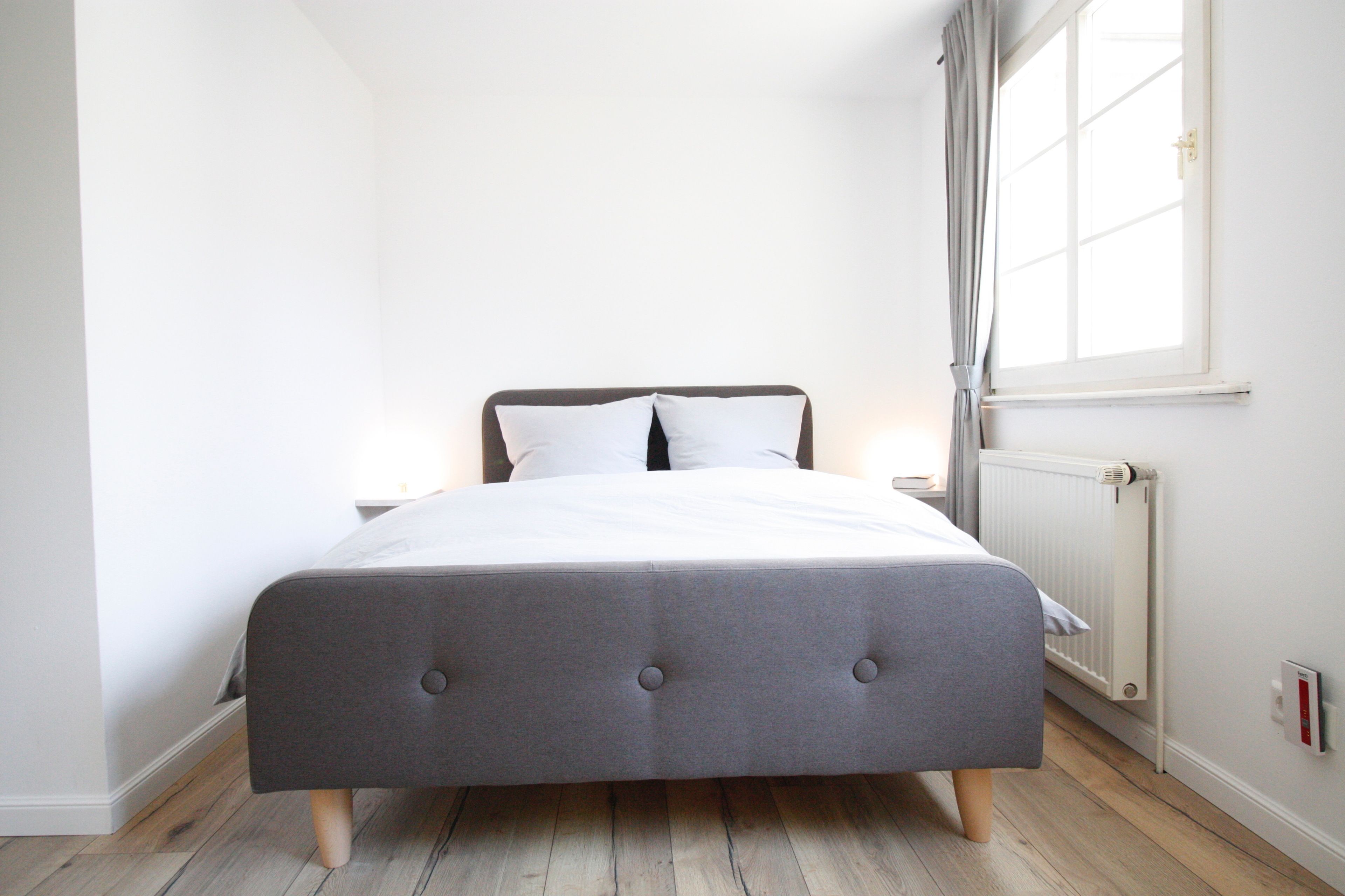 A modern bedroom with a grey upholstered bed, white bedding, a nightstand, a window with curtains, and a radiator.