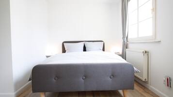 A modern bedroom with a grey upholstered bed, white bedding, a nightstand, a window with curtains, and a radiator.