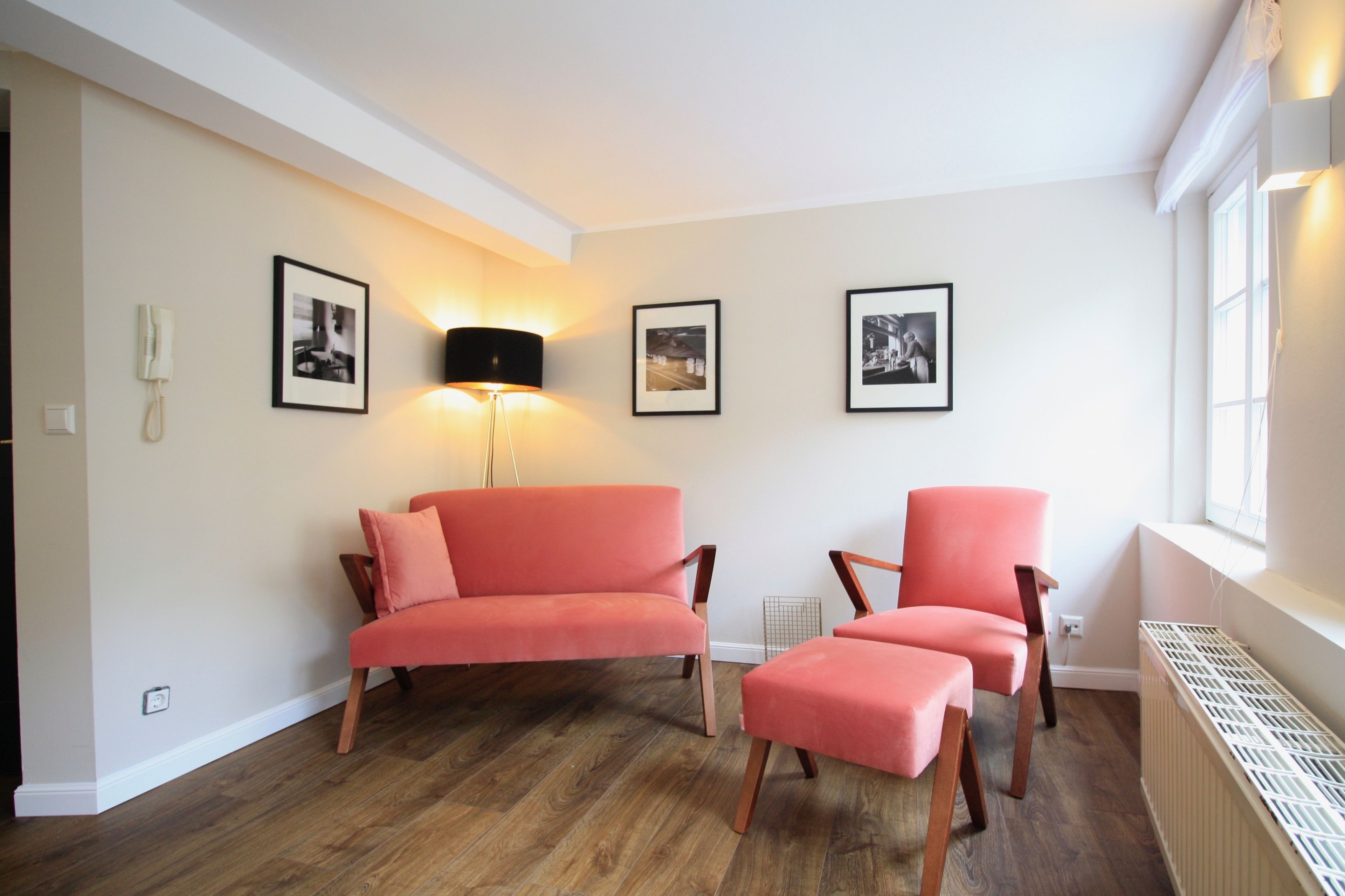 Ein Zimmer mit einer Couch, einem Sessel und einem kleinen Tisch. An den Wänden hängen Bilder in Rahmen und auf dem Boden steht eine Stehlampe.