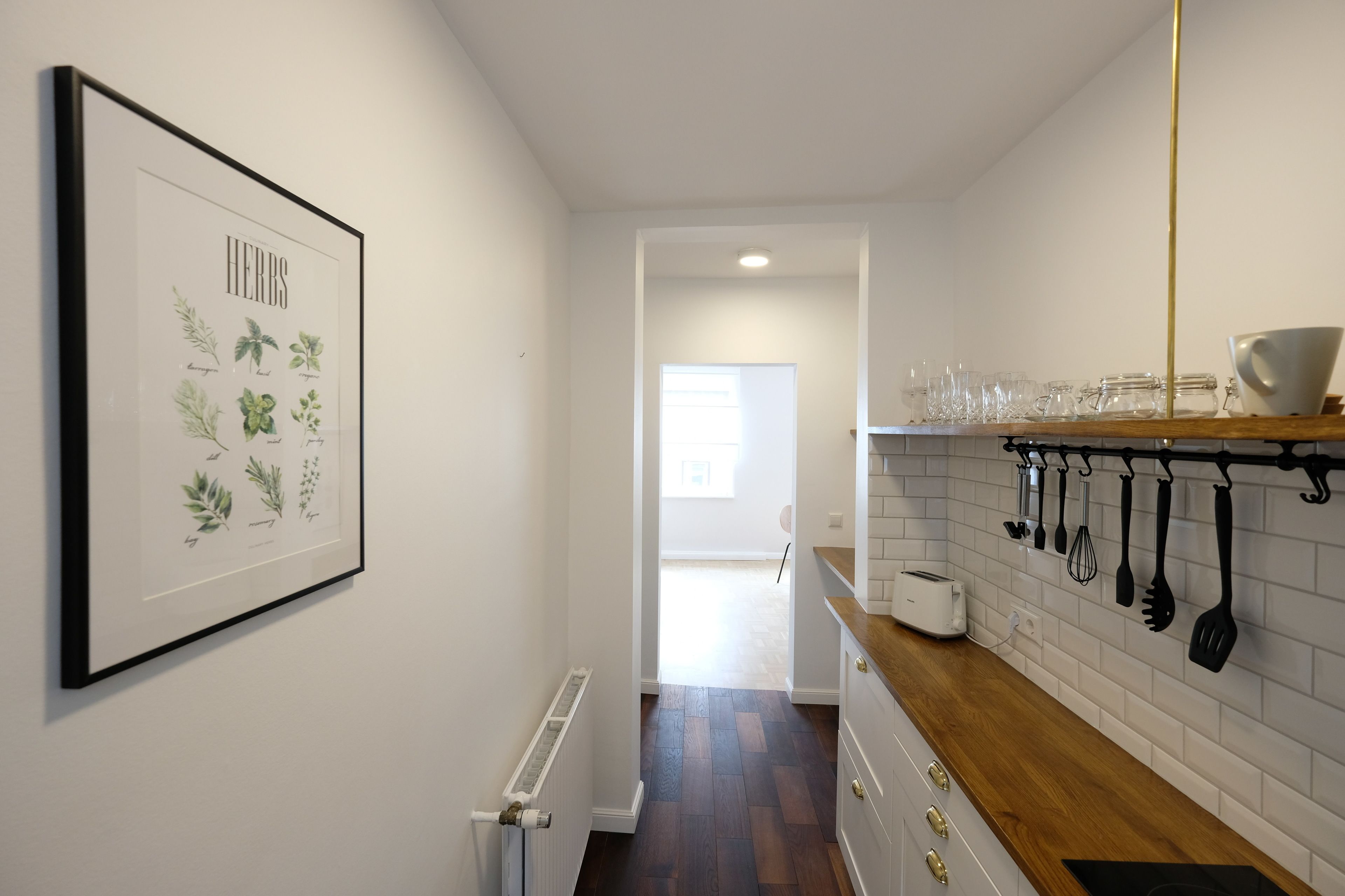 A kitchen with a wooden countertop, white tiled backsplash, and a framed herb illustration on the wall.