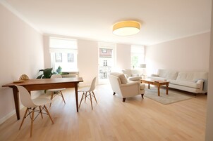 Ampla sala de estar com uma mesa de jantar de madeira, cadeiras brancas, um sofá branco e uma mesa de centro.