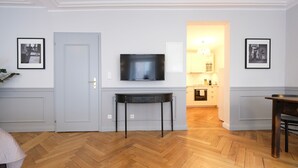 A room with a wooden herringbone floor, a flat-screen TV mounted on the wall, a small table with drawers, and framed pictures on the walls. - Boutique Charme Apartments 14 (Baden-Baden)
