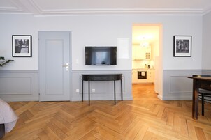 Quarto com piso de madeira em padrão herringbone, televisão de tela plana na parede, mesa pequena com gavetas e quadros nas paredes.