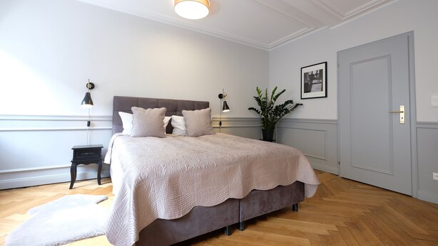 A bedroom with a large bed, bedside tables, a nightstand, a potted plant, and a door. - Boutique Charme Apartments 14 (Baden-Baden)