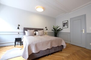 Quarto com cama grande, mesas de cabeceira, um criado-mudo, uma planta em vaso e uma porta.