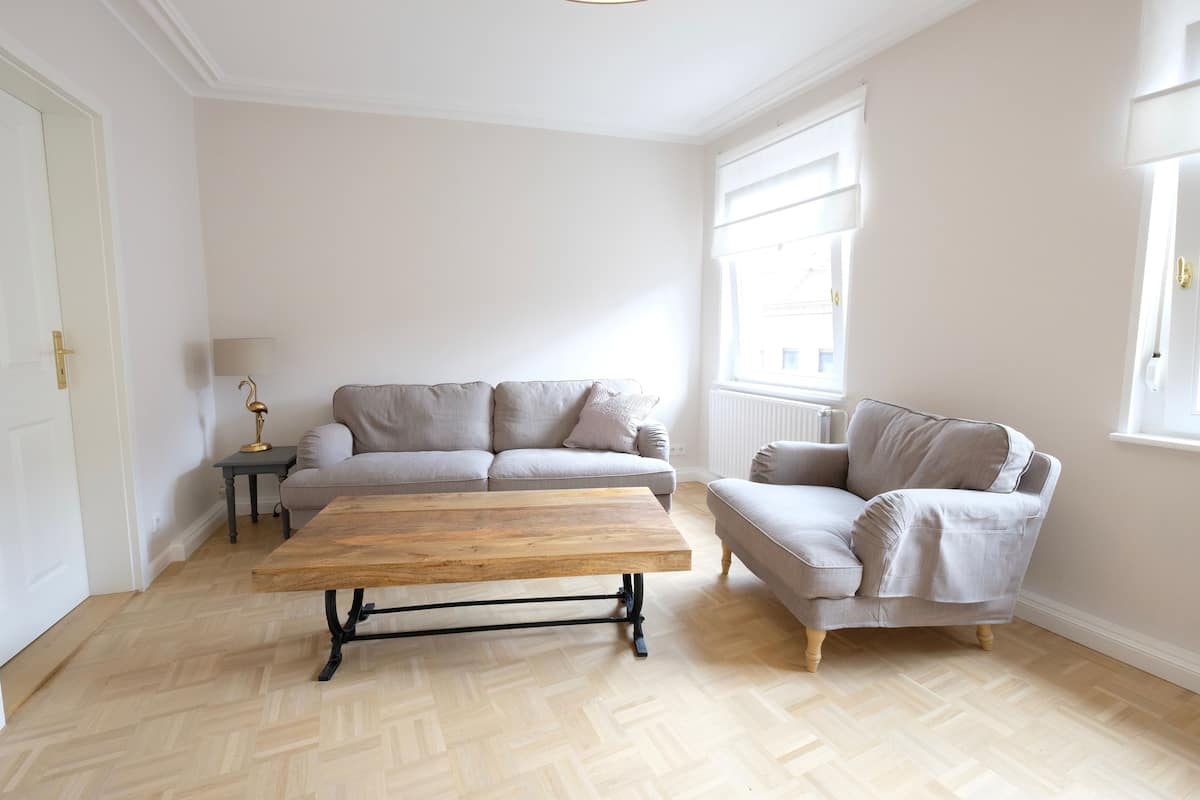 A living room with a wooden coffee table, two grey sofas, a small side table with a lamp, and a window with blinds.