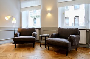 Quarto com duas poltronas, uma mesinha e um abajur. Janelas oferecem vista para prédios.