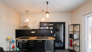 Cocina compacta con encimera negra, electrodomésticos de acero inoxidable y horno integrado. Hay luces colgantes y una pequeña zona de comedor con una silla negra.
