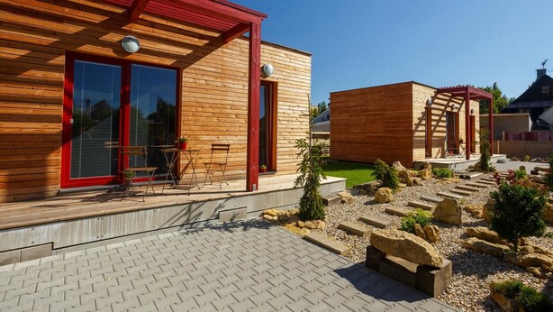 Una casa moderna de madera con techo rojo, ventanas grandes y un patio con mobiliario exterior.