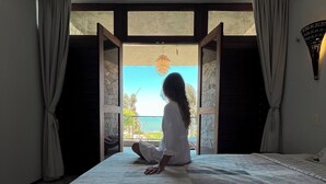 Una habitación con una cama, una persona sentada en el suelo y vistas a través de una puerta abierta.