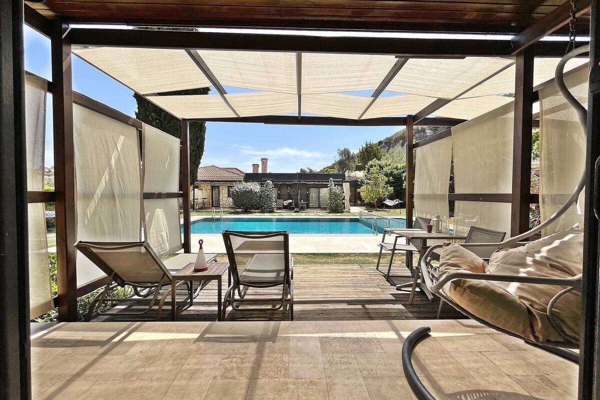An outdoor patio with a pool, covered seating, and a view of a house in the background.