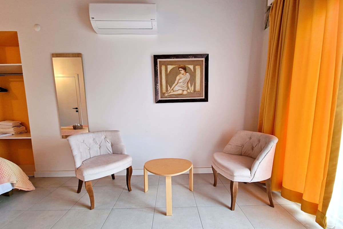 A hotel room with two armchairs, a small round table, a mirror, and an air conditioning unit on the wall.