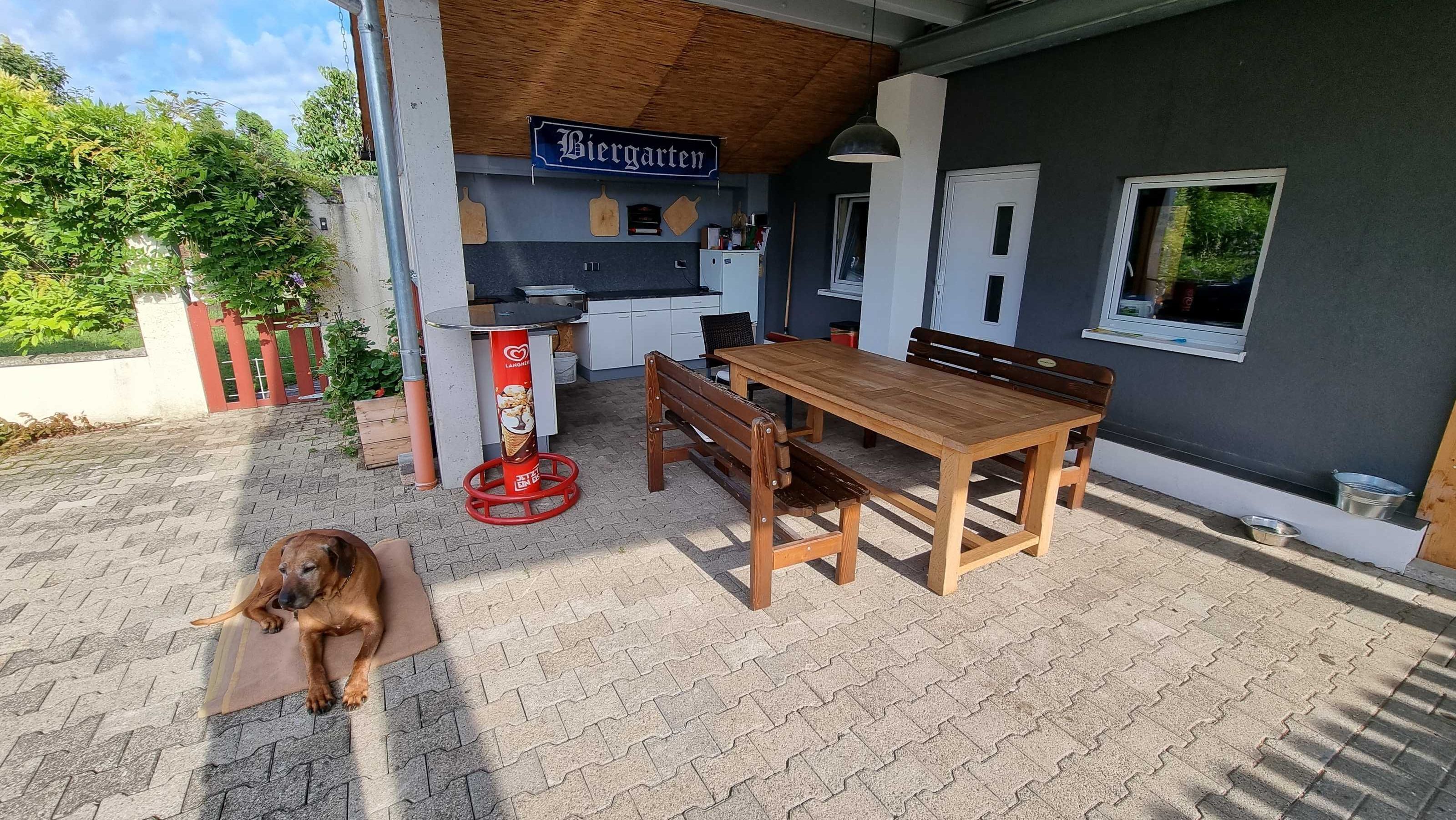 En utesittplats med ett träbord och bänkar, en hund som ligger på marken och en skylt med texten ”Biergarten”.