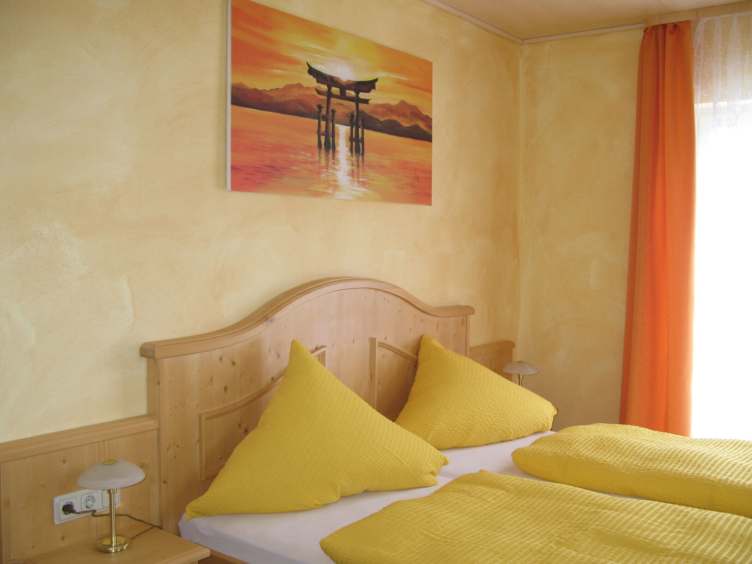 Una stanza con un letto, una testata in legno, cuscini gialli, un quadro raffigurante un tramonto e una tenda arancione.