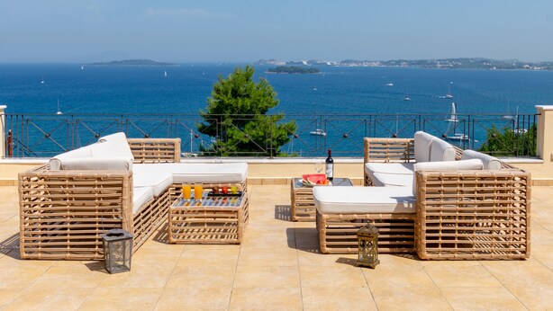 A terrace with wicker furniture, a glass table with a bottle and fruit, and a view of the sea with boats. - Villa Kommeno (Corfu)
