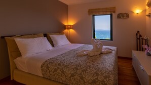 A neatly made bed with a floral bedspread and white pillows, a bedside lamp, a window with a view of the sea, and a small desk with a vase of flowers. - Villa Kommeno (Corfu)