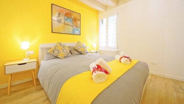 A bright yellow room with a bed, a nightstand, a lamp, and a painting on the wall. - DolceVita Apartments N. 443 (Venice)
