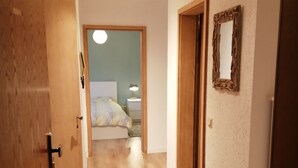 Ein Flur mit Holzboden, einem Bett mit weißem Kopfteil, einem Nachttisch mit Lampe und einem an der Wand befestigten Spiegel.