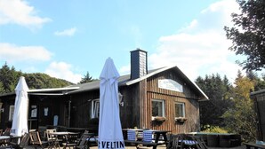 Ein Außenbereich mit Holztischen und -stühlen, einem weißen Sonnenschirm und einem Holzhaus mit Schornstein.