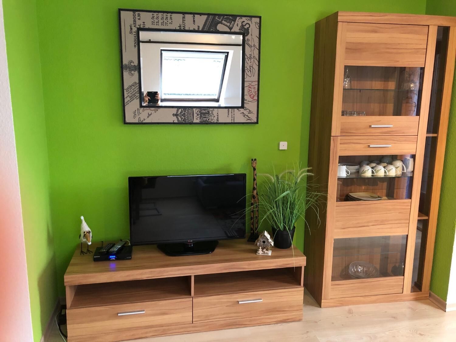 A room with a green wall, a wooden entertainment unit with a flat-screen TV, a small plant, and a wooden cabinet with glass doors.
