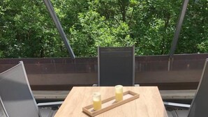 Ein quadratischer Holztisch mit zwei Gläsern und einer Tablett, umgeben von vier Metallstühlen, draußen mit Blick auf Bäume.