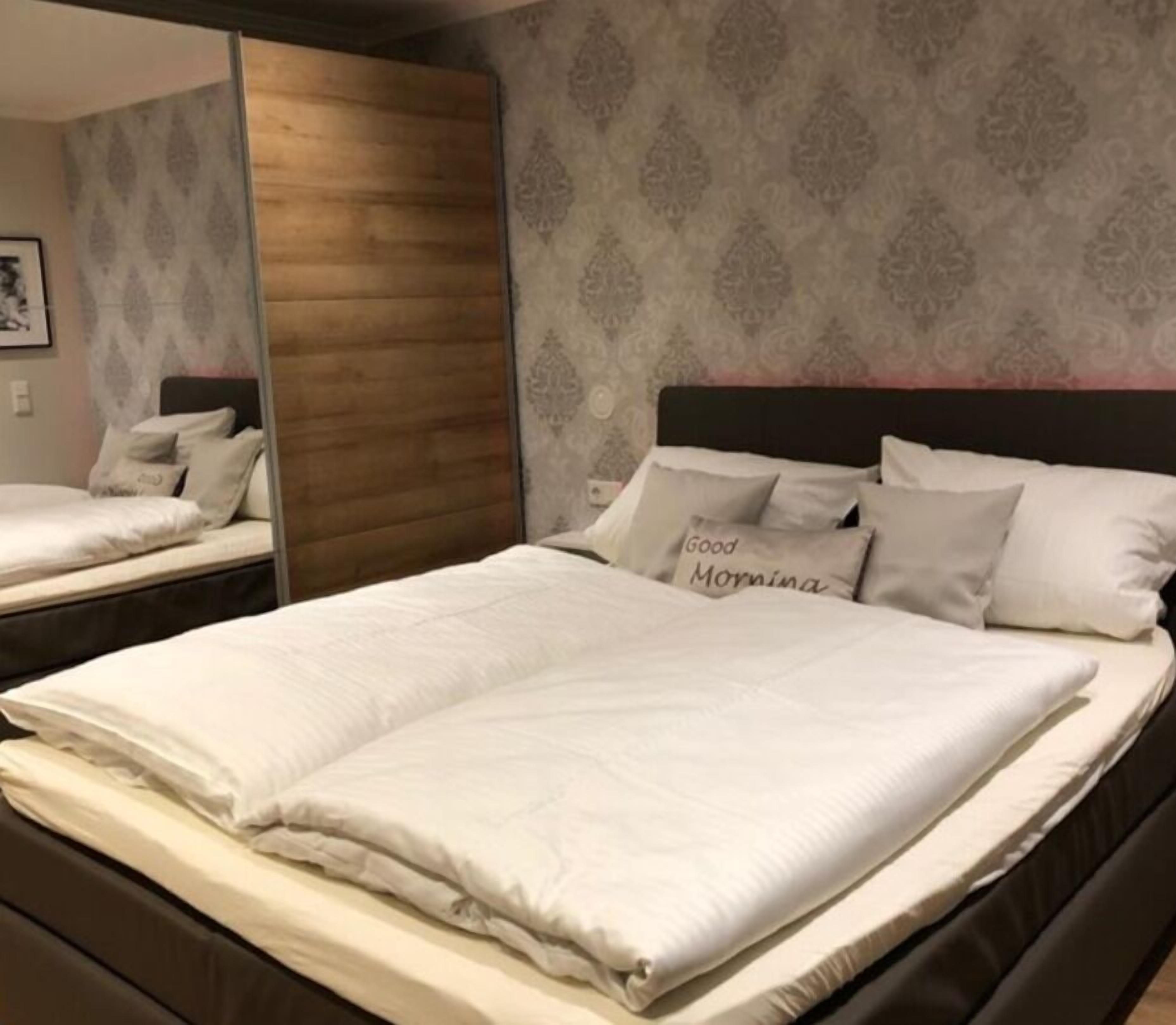 A neatly made bed with white linens and pillows, a dark headboard, and a patterned wallpaper.