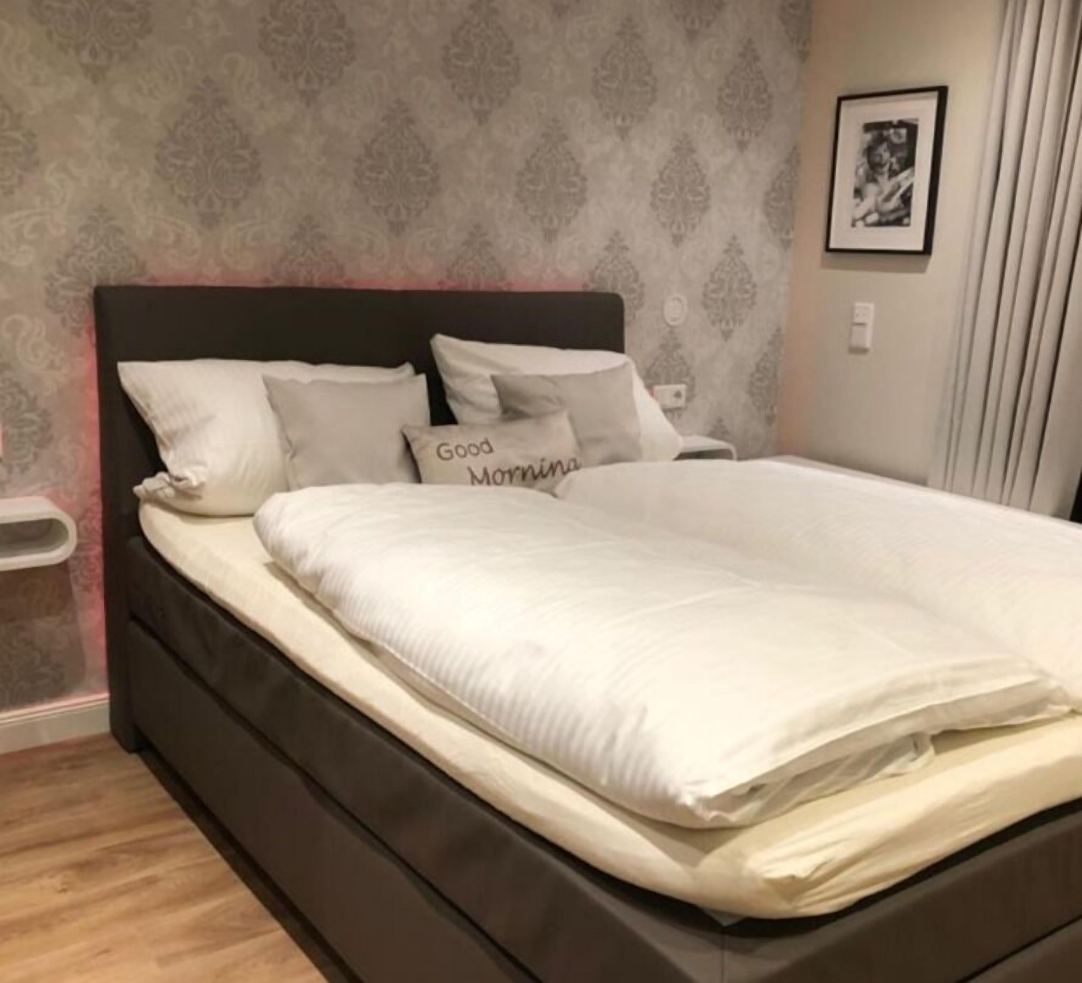 Un lit bien fait, avec du linge de lit blanc et des oreillers, dont l’un porte l’inscription « Bonjour ».
