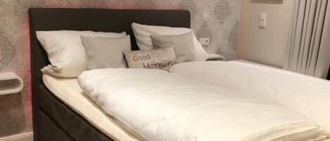Un lit bien fait, avec du linge de lit blanc et des oreillers, dont l’un porte l’inscription « Bonjour ».