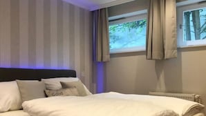 Ein ordentlich bezogenes Bett mit weißen Leinen und Kissen, ein dunkles Kopfbrett und ein Fenster mit Vorhängen.