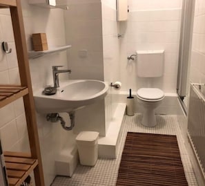 A small, clean bathroom with a white sink, toilet, and shower. There are wooden shelves above the sink and a small trash bin next to the toilet.