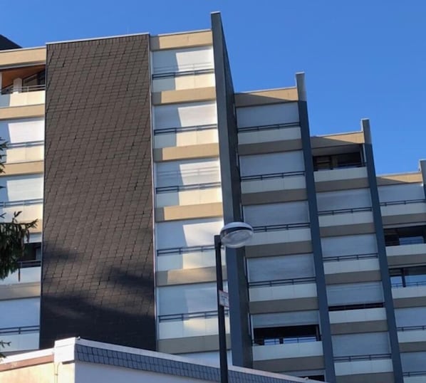 A multi-story building with a modern architectural design, featuring a prominent vertical element on the left side and a series of balconies on the right.
