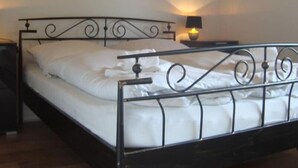 Una cama con estructura metálica y ropa de cama blanca, una mesita de noche con una lámpara y un techo de madera.