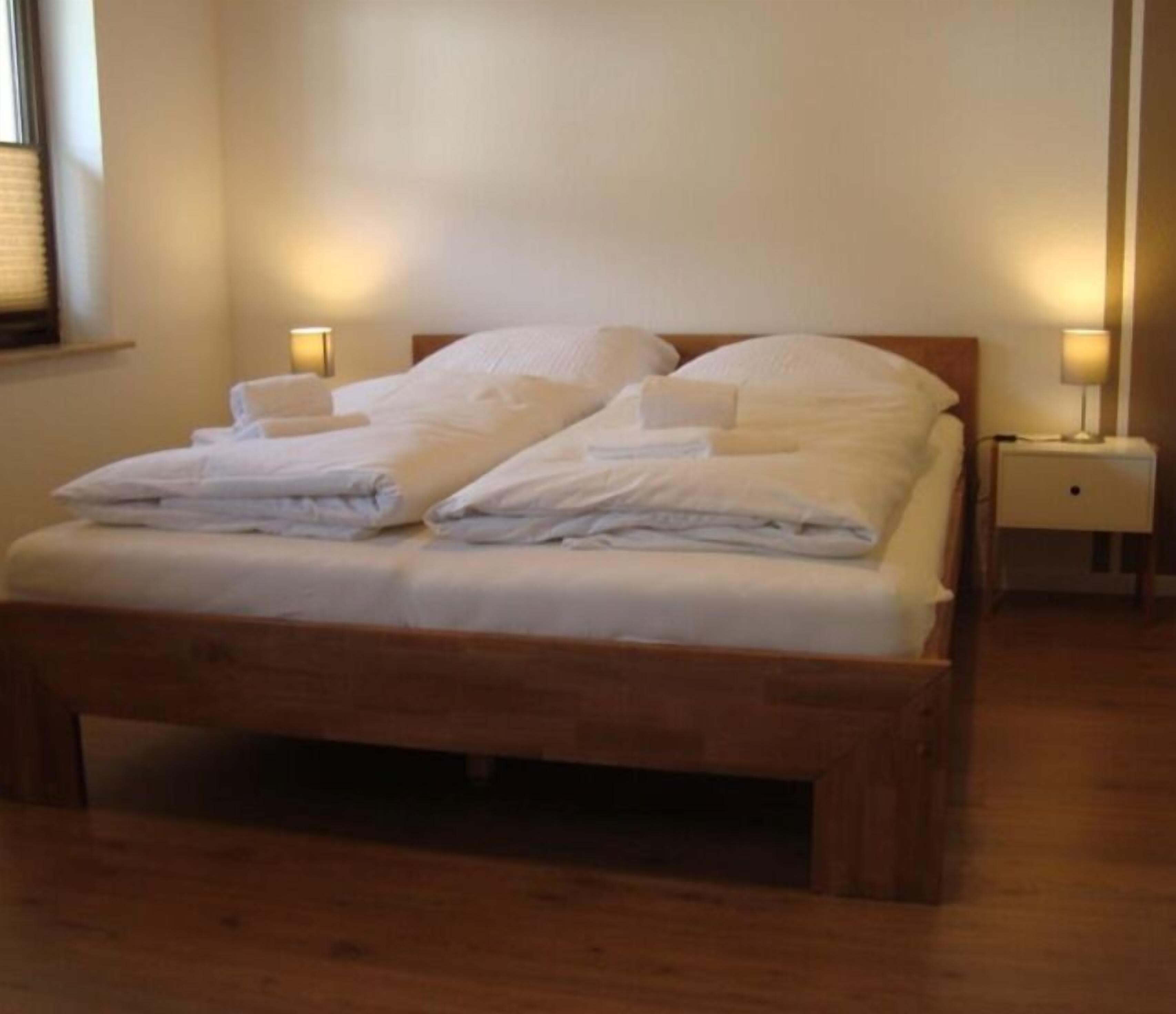 Ein ordentlich bezogenes Bett mit weißen Leinen und Kissen, ein hölzerner Kopfteil und zwei Nachttische mit Holz und jeweils einer Nachttischlampe.