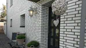 A building with white brick walls, a black door, and a heart-shaped decoration. There are two wicker baskets with plants on the steps and a bench outside. - Apartment Mühlenberg (Winterberg)