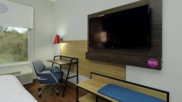 A hotel room with a bed, a desk, a chair, a TV mounted on the wall, and a window with blinds.