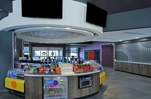 A modern, well-lit food court with a variety of food and drink options, including a vending machine and a self-service beverage dispenser.