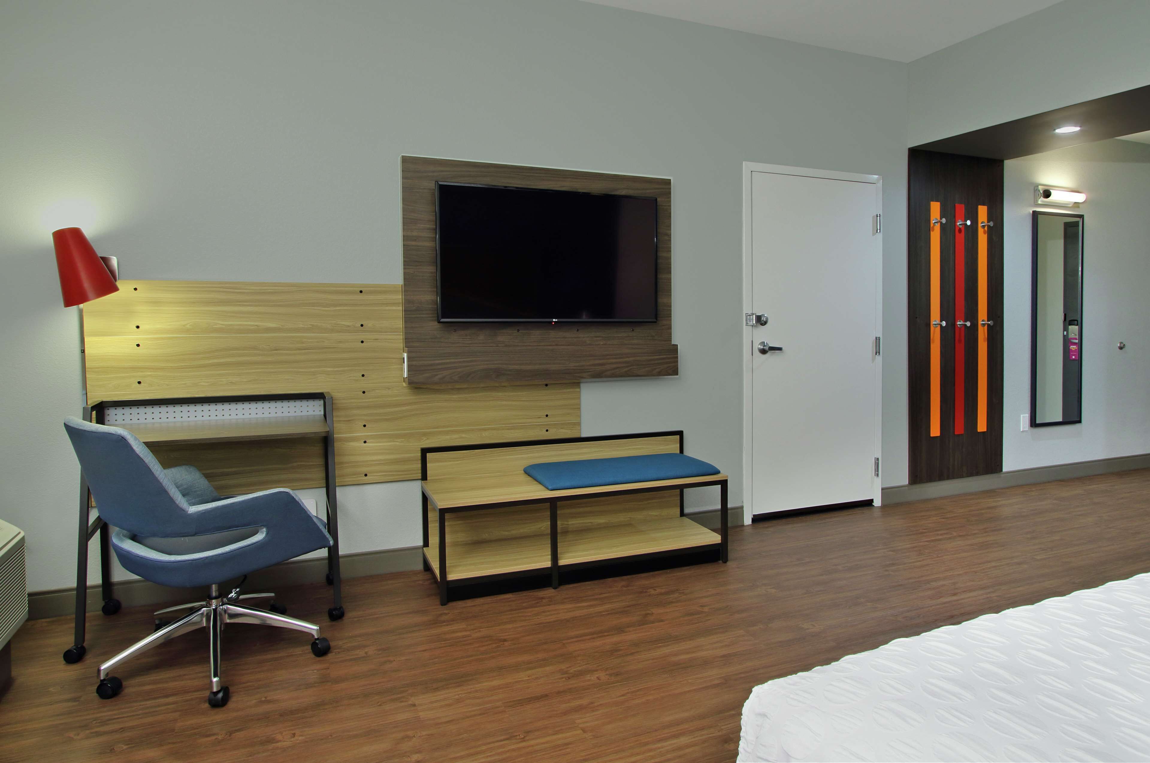 Quarto de hotel com escrivaninha de madeira, uma cadeira azul, televisão na parede, um banco e uma porta com espelho.