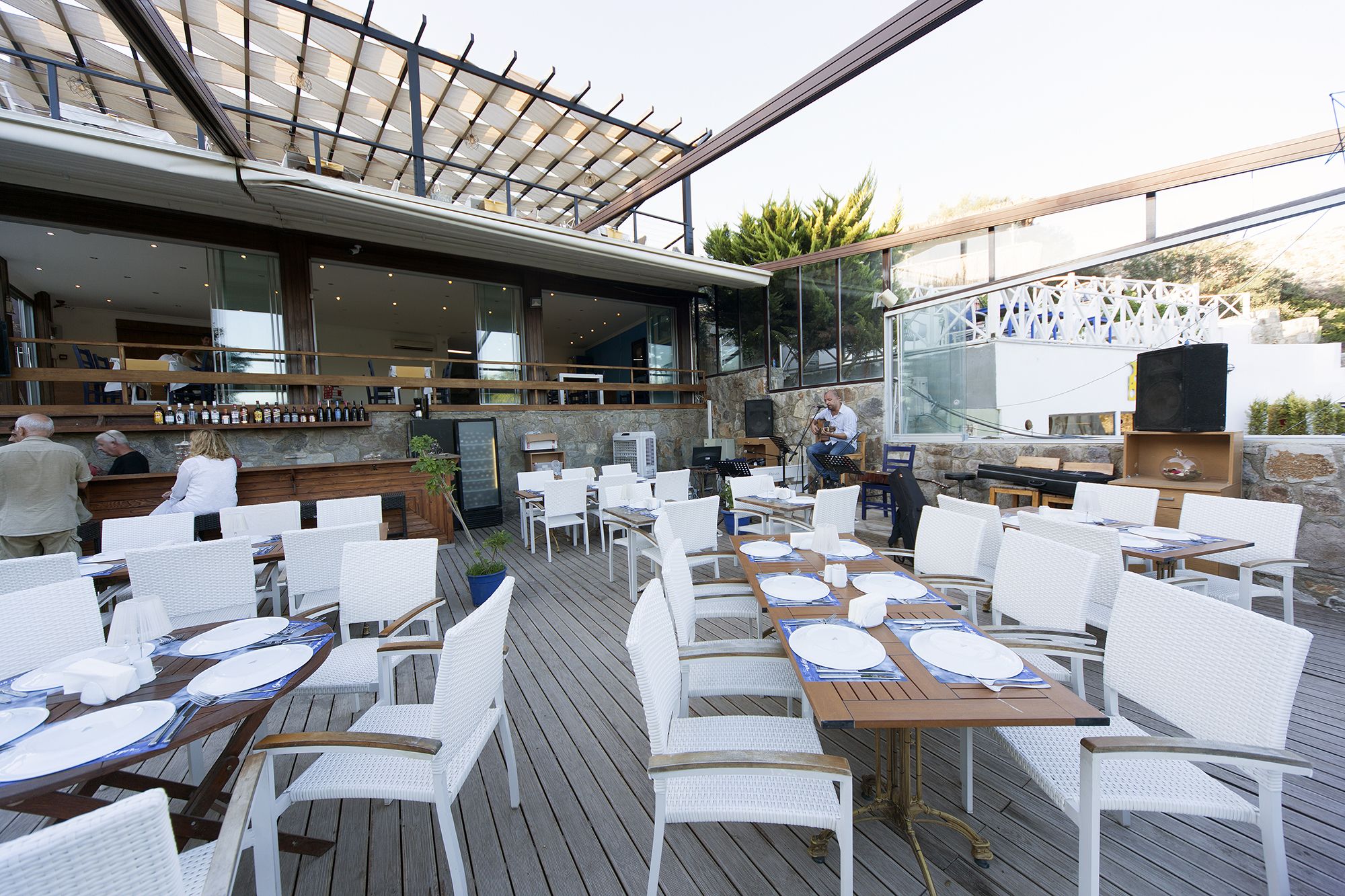 An outdoor dining area with white wicker chairs and tables, a bar area with bottles, and a musician performing.
