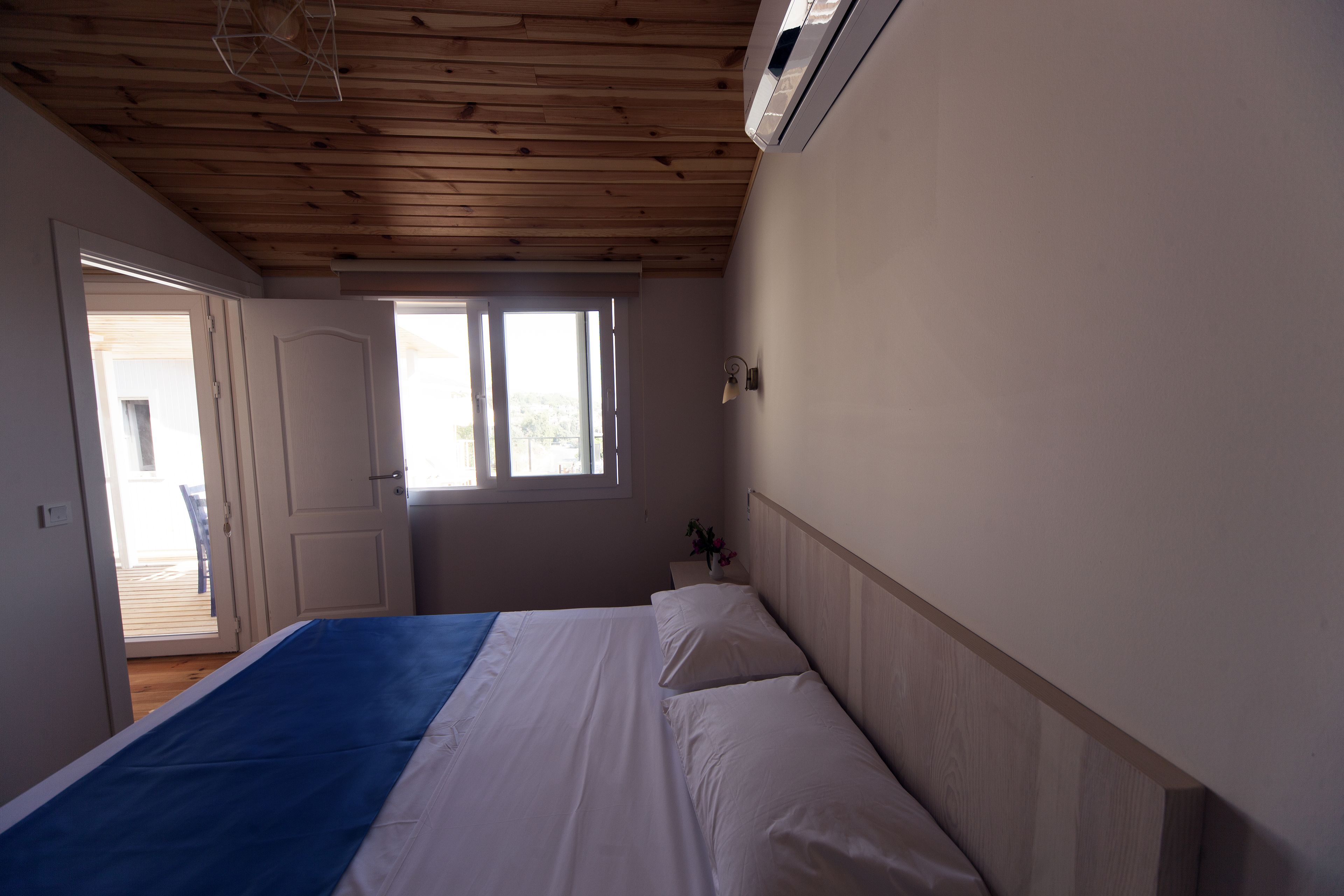 A bedroom with a wooden ceiling, a bed with white linens, a blue accent on the bedspread, a nightstand with a lamp, and a window with a view of the outside.