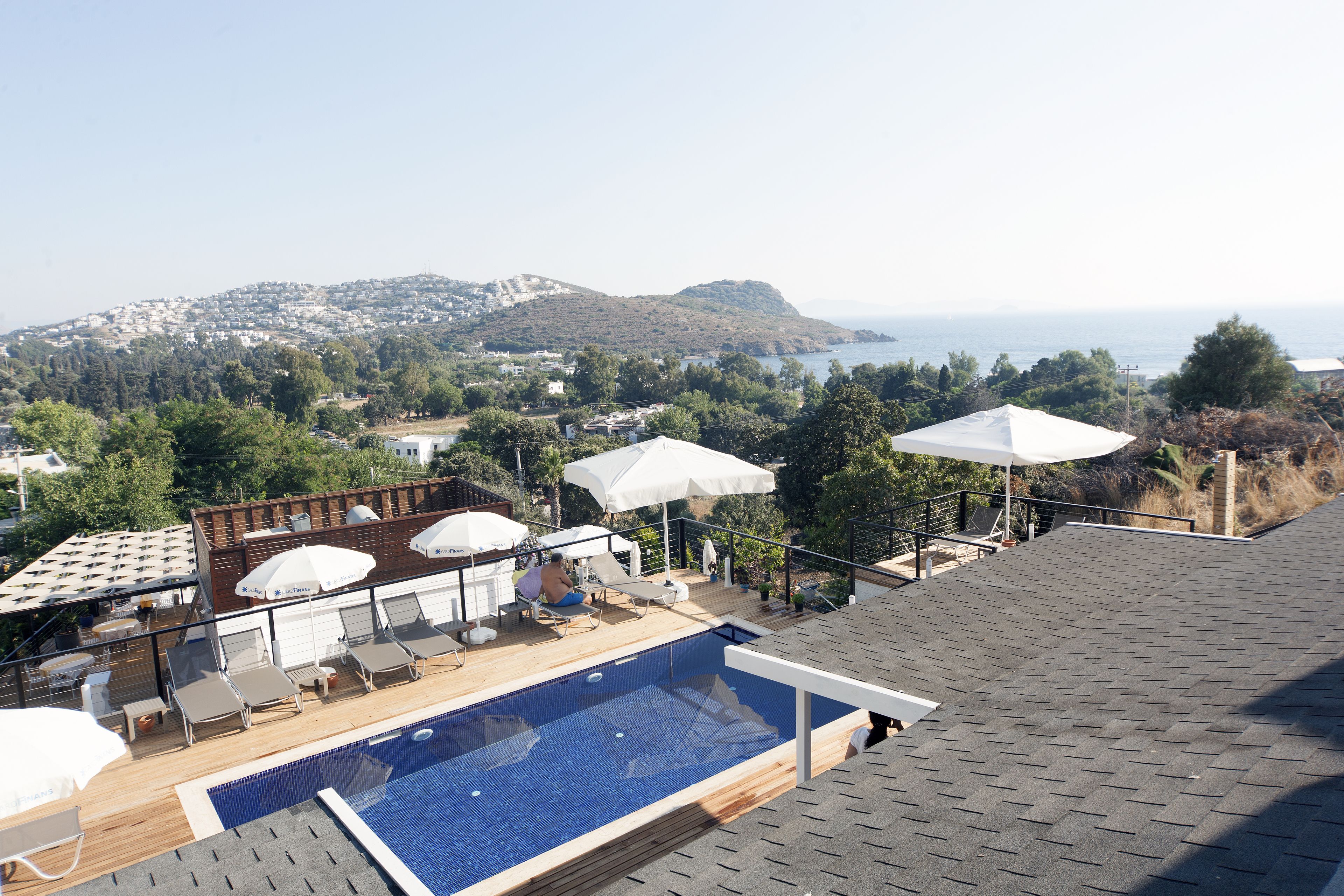 Piscina en la azotea con sillas de descanso y sombrillas, con vistas a un pueblo costero y colinas.