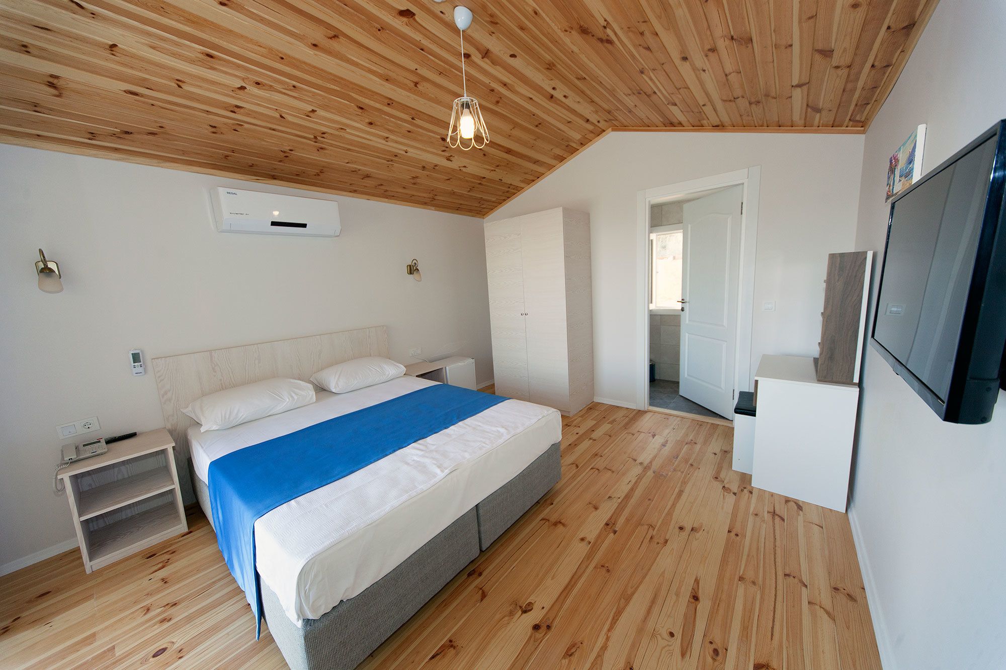 Slaapkamer met houten plafond, bed met blauwe deken, nachtkastje, televisie en garderobe.