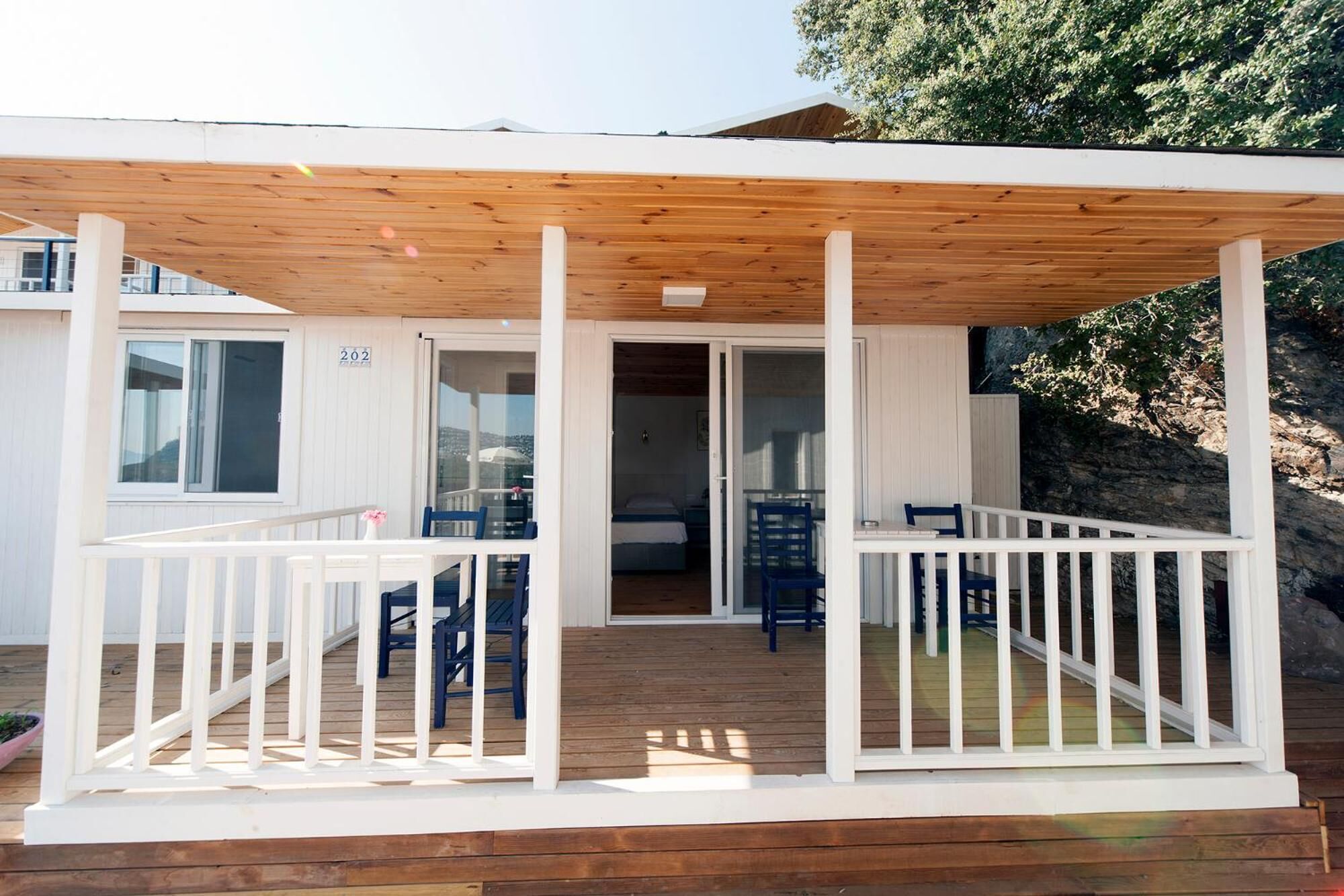 A covered outdoor area with a wooden deck, white railings, and a glass door leading to a room with a bed and a chair.