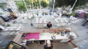 An outdoor dining area with multiple tables and chairs, a bar area with bottles and glasses, and a view of the street and parked cars. - Metin's Gumusluk (Bodrum)