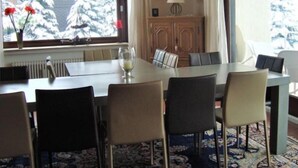 A dining area with a table set for six, a radiator, a cabinet with a lamp, and a view of snow-covered trees through a window. - Ferienhaus Beja (Winterberg)