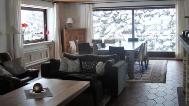 A cozy living room with a sofa set, a dining table, and a view of snowy trees through large windows. - Ferienhaus Beja (Winterberg)