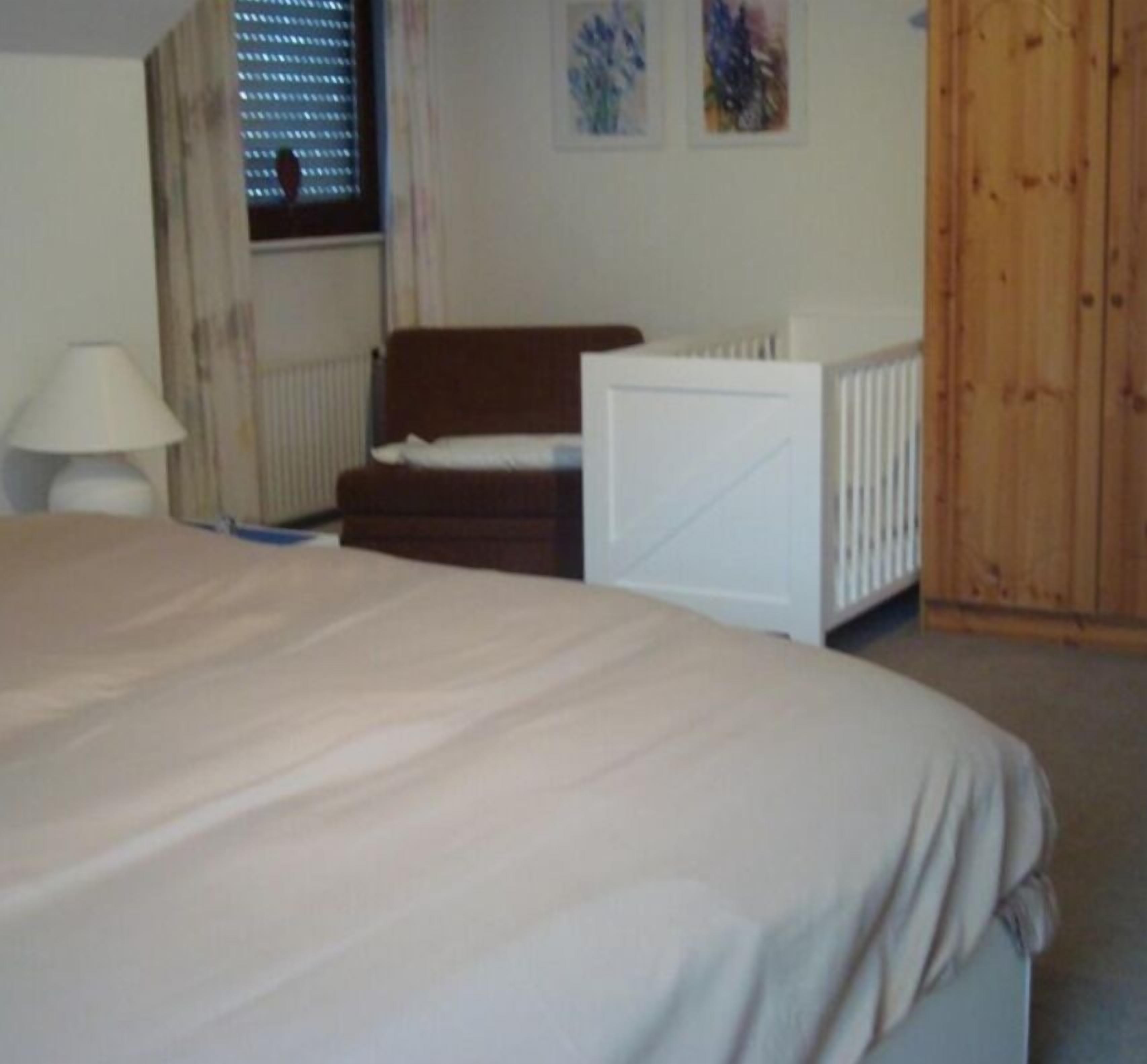 A bedroom with a large bed, a wooden chair, a white bedside table, and a window with blinds.